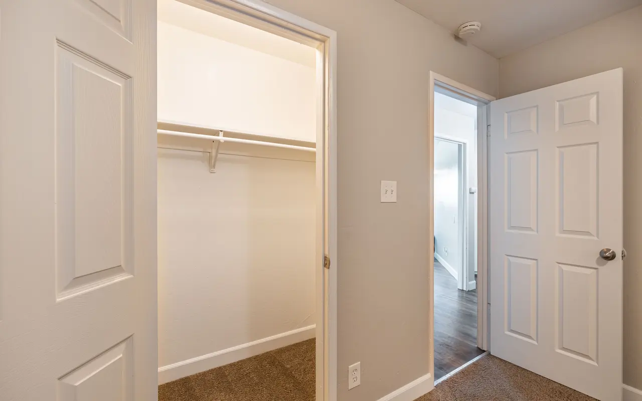 A view of a small hallway featuring two doors; one leads to a closet with a hanging rod, and the other leads to an adjoining room. The walls are painted a light color, and the flooring appears to be carpeted in a tan hue.