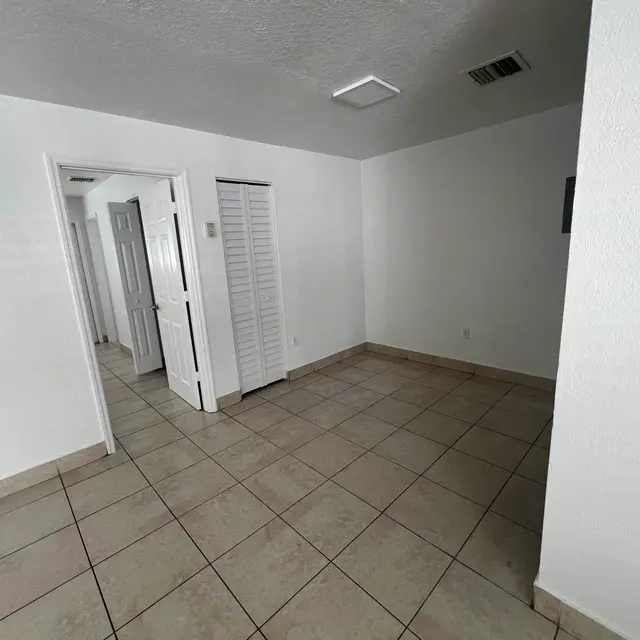 A spacious, empty room with light-colored tile flooring, white walls, and multiple doorways leading to other areas.