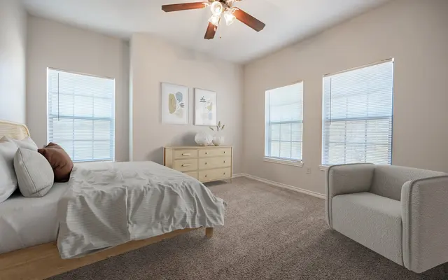 A modern bedroom featuring a large bed, a light-colored dresser, and a cozy armchair. The room has multiple windows allowing natural light to illuminate the space, with a neutral color palette and soft textures throughout.