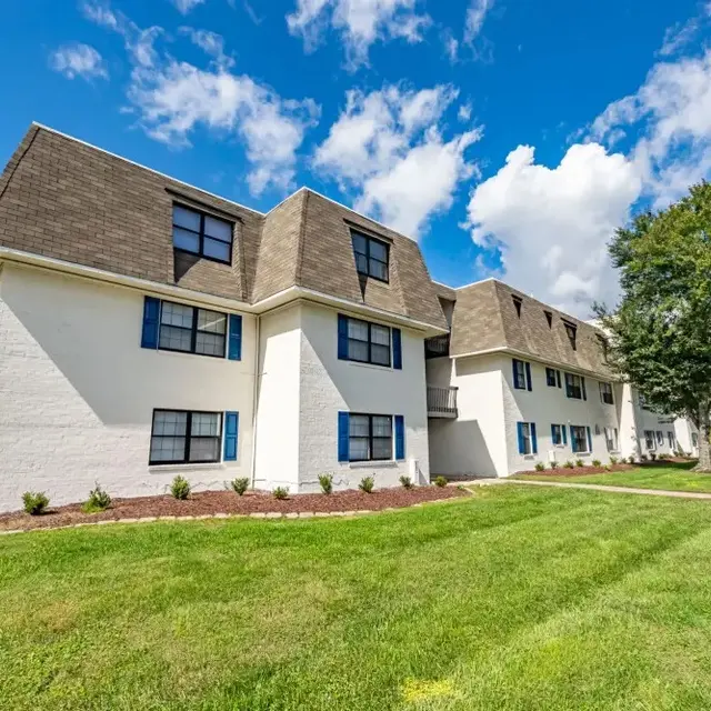 A multi-story apartment building with a unique roof design, featuring white walls and blue window shutters, situated on a green lawn under a partly cloudy sky.
