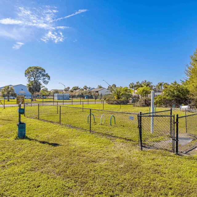 Spacious Dog Park A spacious dog park enclosed by a black fence, featuring agility equipment and benches. The area has green grass, trees, and clear blue skies in the background.