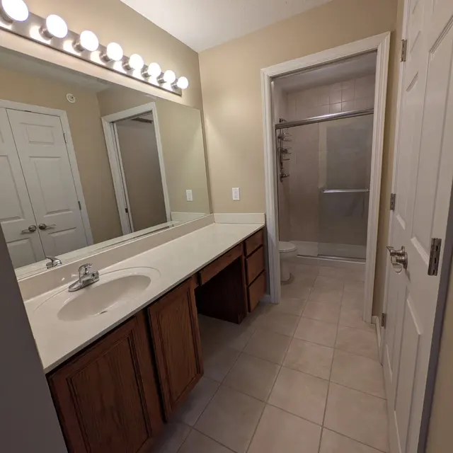 A well-lit bathroom featuring a double sink vanity with wooden cabinetry, a large mirror with bright lights above, beige walls, and tile flooring leading to a shower area.
