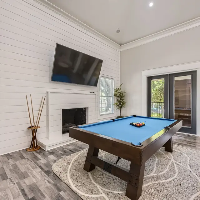 A modern recreational room featuring a blue pool table, a large flat-screen TV mounted on a white shiplap wall, and glass doors leading outside. There are decorative plants and a cozy rug on the floor.
