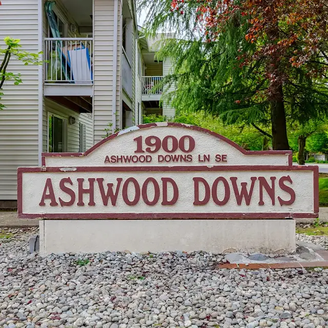 A sign that reads 'ASHWOOD DOWNS' with the address '1900 ASHWOOD DOWNS LN SE' below it, situated in front of a residential building surrounded by greenery.
