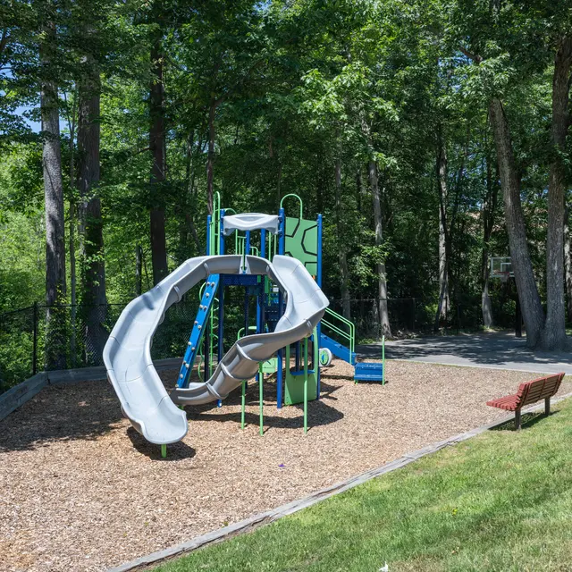 A playground featuring a gray and green slide surrounded by mulch and benches. It is set in a wooded area with trees in the background.
