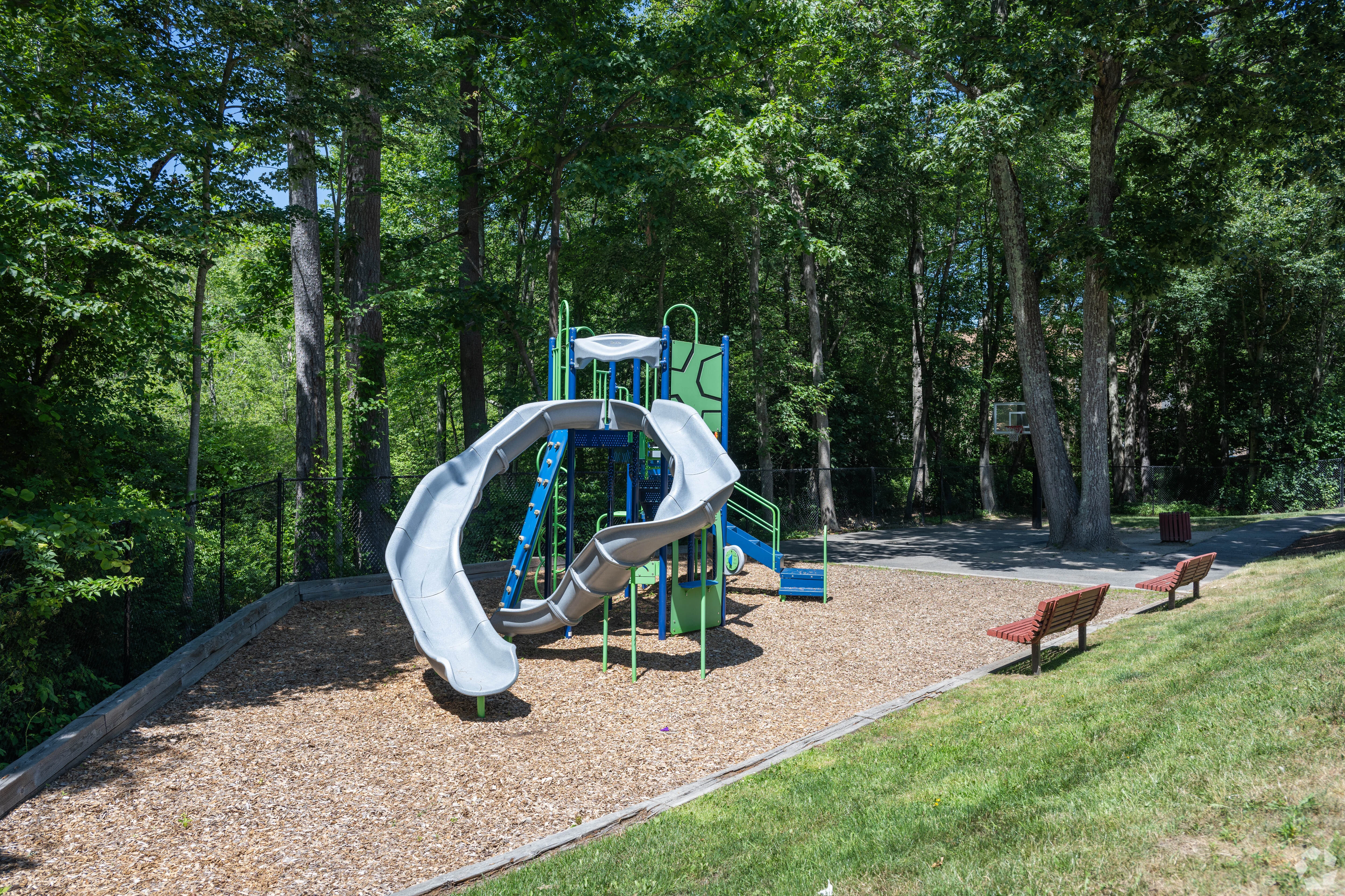 Children's Playground with Slide A playground featuring a gray and green slide surrounded by mulch and benches. It is set in a wooded area with trees in the background.