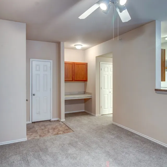 Interior View of Small Apartment Living Room An interior view of a small apartment living room with beige walls and carpet. It features a ceiling fan, white doors, and a small kitchenette visible in the background.