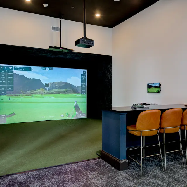 An indoor golf simulator setup featuring a large projection screen showing a golf course, with a tee area and a bar counter with stools nearby.