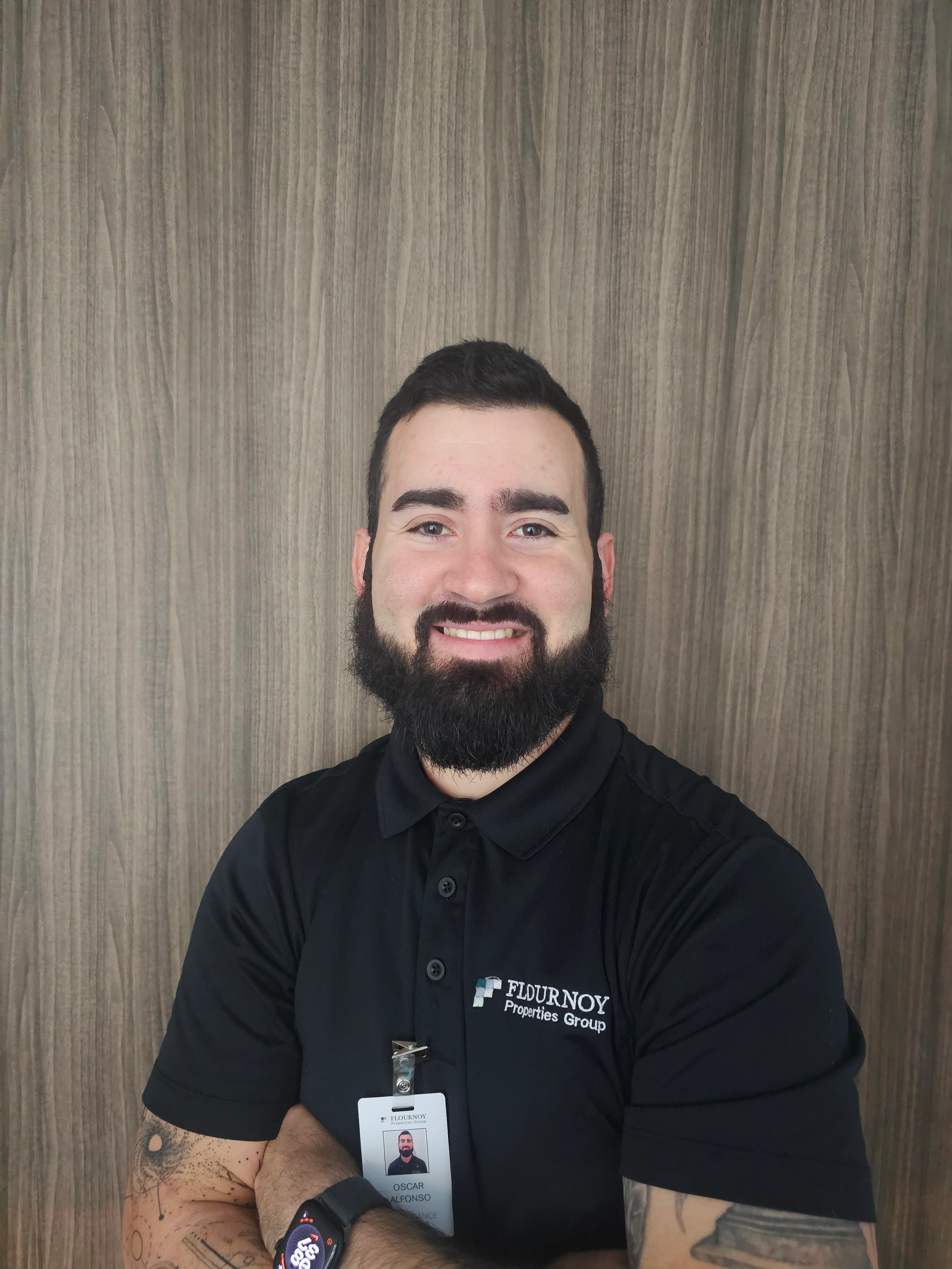 Confident Man in Black Polo Shirt A smiling man with a beard wearing a black polo shirt stands against a wooden wall, arms crossed.