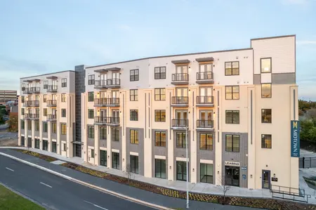 A modern apartment building with five levels, featuring a blend of white and grey exterior materials, multiple balconies, and large windows, alongside a paved roadway.