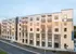 A modern apartment building with five levels, featuring a blend of white and grey exterior materials, multiple balconies, and large windows, alongside a paved roadway.