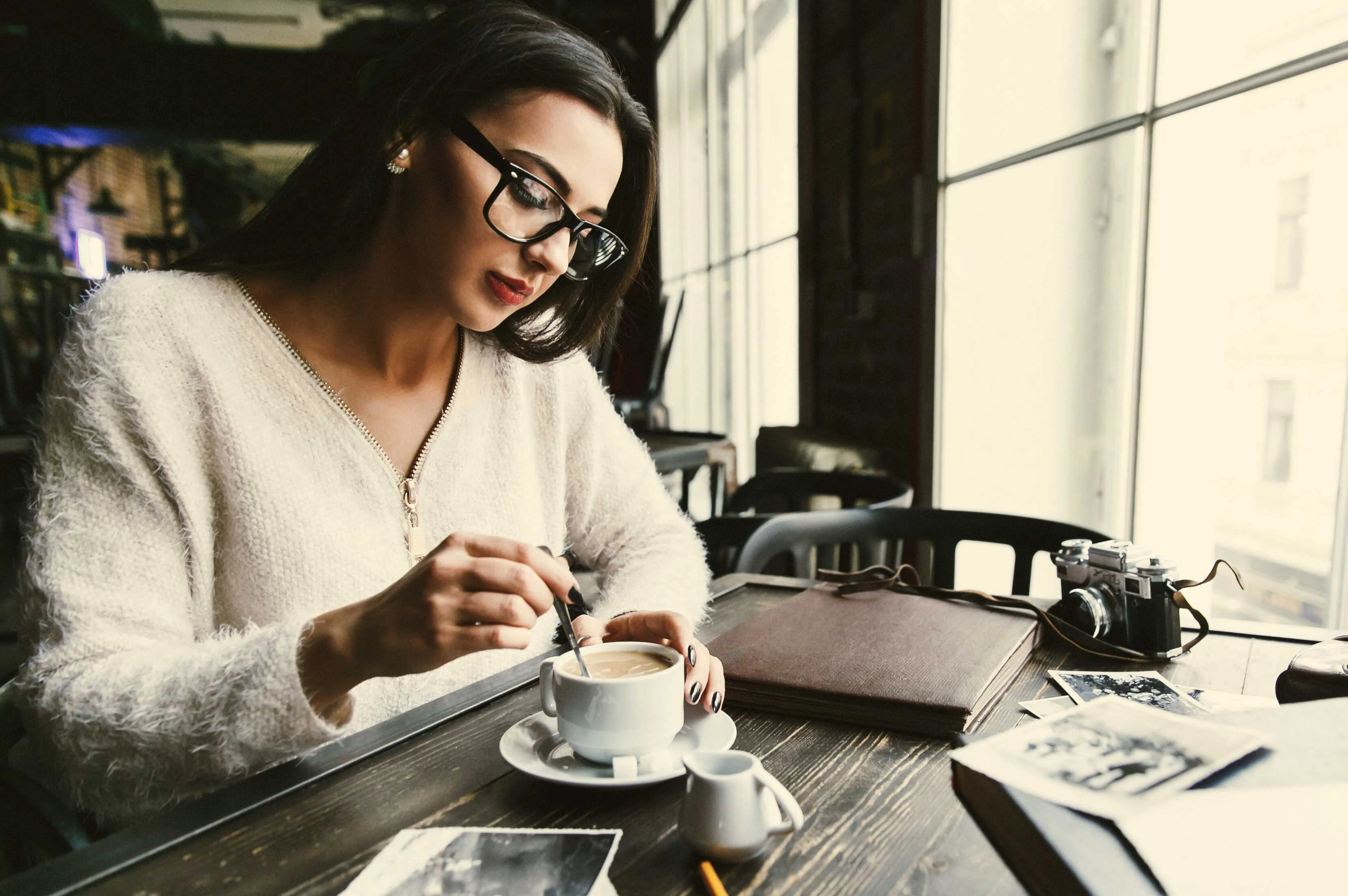 Coffee Break in a Cozy Café A woman with glasses enjoys coffee at a table in a café, stirring her drink while sitting next to a notebook and photographs.