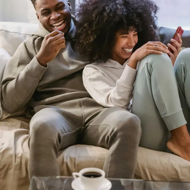 A man and woman sitting together on a couch, both laughing while looking at a phone. The man has short hair and is wearing a gray hoodie and pants. The woman has curly hair and is dressed in a light hoodie and pants. A cup of coffee sits on a glass table in front of them.