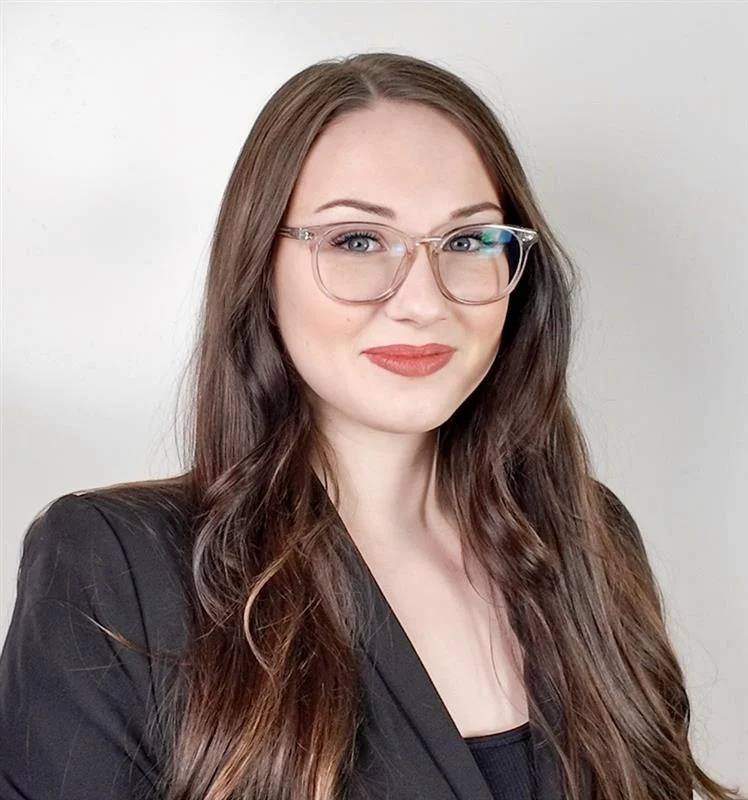 Professional Portrait A woman with long dark hair and glasses wearing a black blazer, smiling against a plain background.