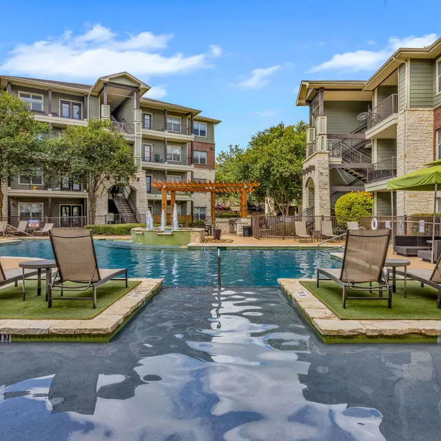 A vibrant apartment pool area featuring a large pool surrounded by lounge chairs and greenery, with modern apartment buildings in the background.