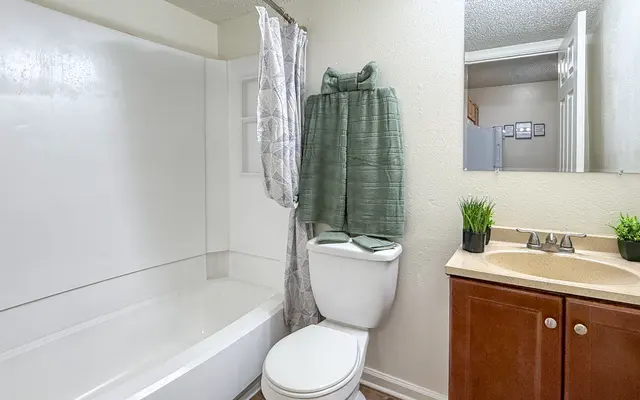 A clean and modern bathroom featuring a white bathtub, a shower curtain, a toilet, and a wooden vanity with a sink. A towel hangs on the wall, and small green plants add a touch of decor.