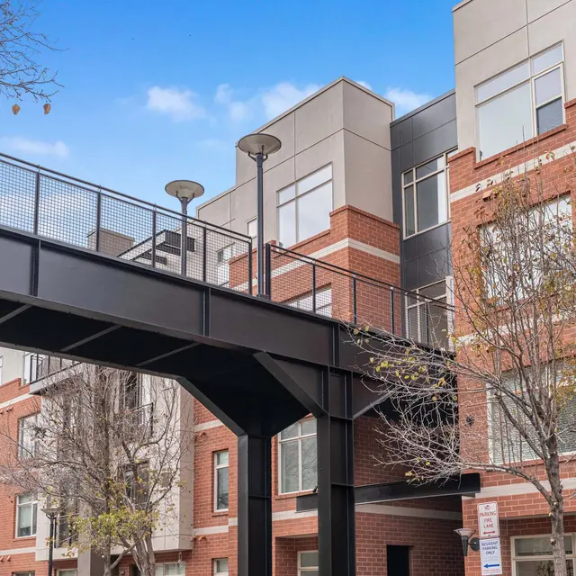 A modern apartment complex featuring multiple floors, large windows, and a pedestrian bridge connecting two sections, with trees nearby.