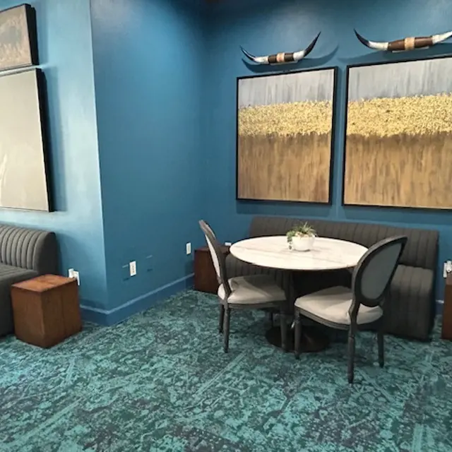 A modern interior space featuring a teal wall, two abstract paintings, a round dining table with a small plant, and a sofa with wooden side tables.
