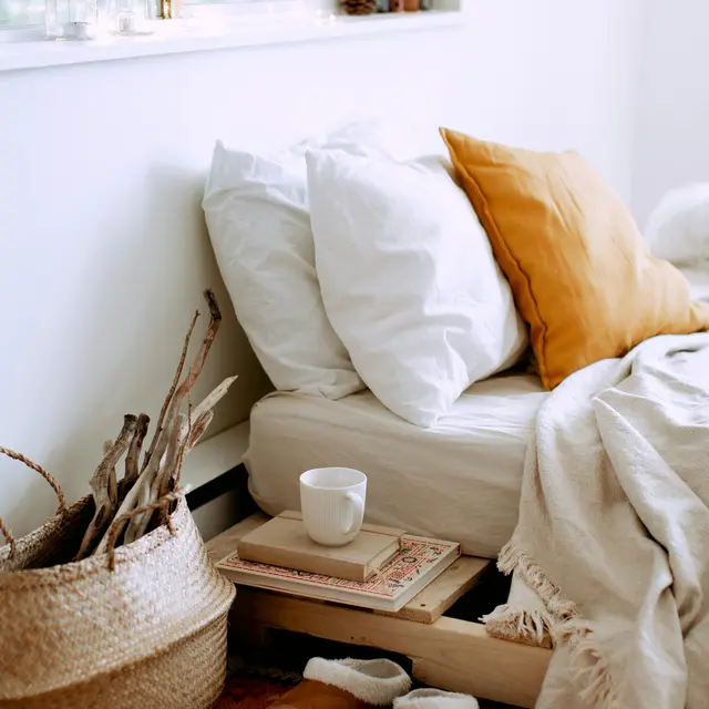 Cozy Bedroom Corner A cozy bedroom corner featuring a bed with white pillows and a mustard-colored throw pillow. Nearby, there is a small wooden side table with books and a cup. A woven basket with sticks is next to the bed, and slippers are placed on a patterned rug.