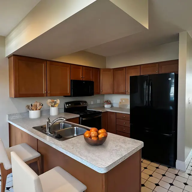 A modern kitchen featuring wooden cabinets, a black refrigerator, and a central island with a bowl of oranges. The kitchen has a tiled floor and is connected to a living area with a light-colored couch.