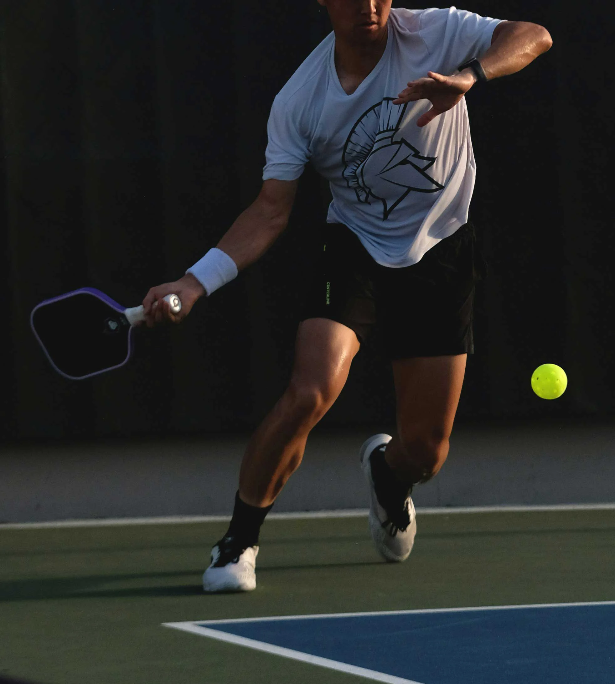 Tennis Player in Action A male player in a white t-shirt and black shorts is about to hit a yellow tennis ball with a paddle on a court.