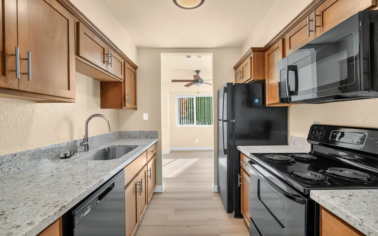 A modern kitchen featuring wooden cabinets, granite countertops, and stainless steel appliances, including a dishwasher, stove, and microwave. A doorway leads to a bright living space with a ceiling fan and natural light coming through a window.