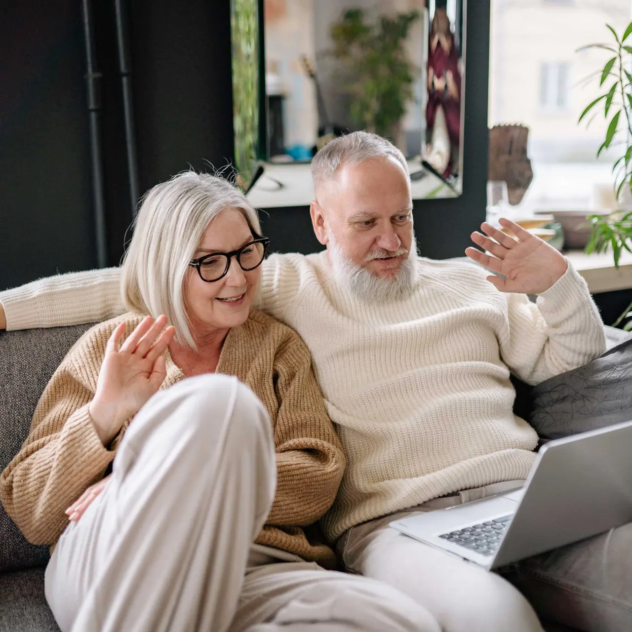 Elderly Couple Video Calling An elderly couple sitting on a couch, smiling and waving at a laptop during a video call. They are surrounded by indoor plants and a cozy living room atmosphere.
