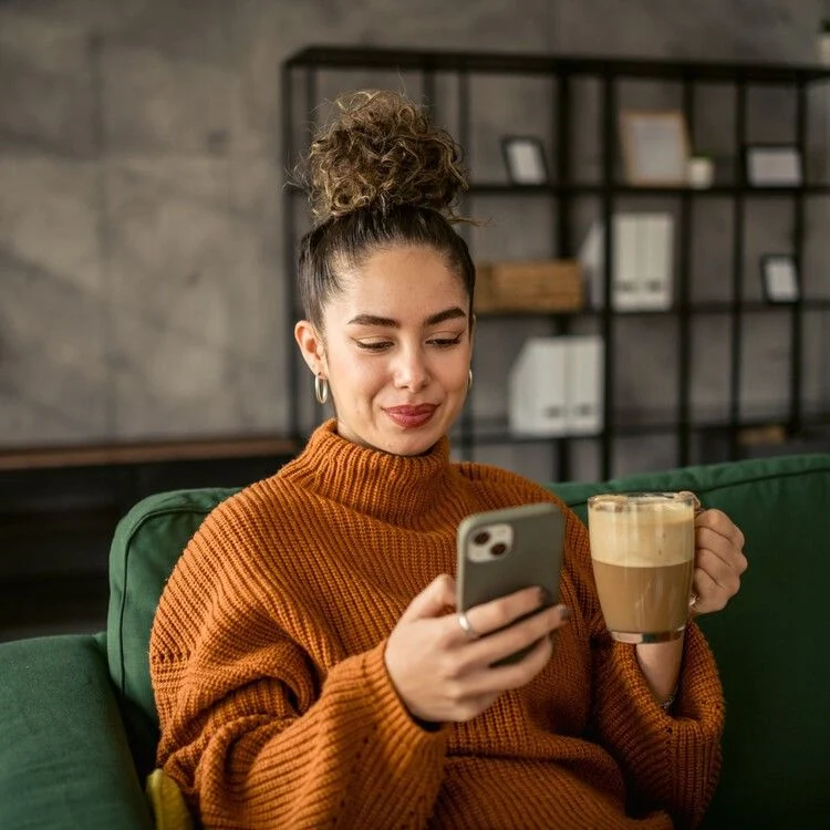 Cozy Coffee Moment A woman sitting comfortably on a green couch, holding a smartphone in one hand and a cup of coffee in the other, wearing a cozy orange sweater, with a content expression on her face.