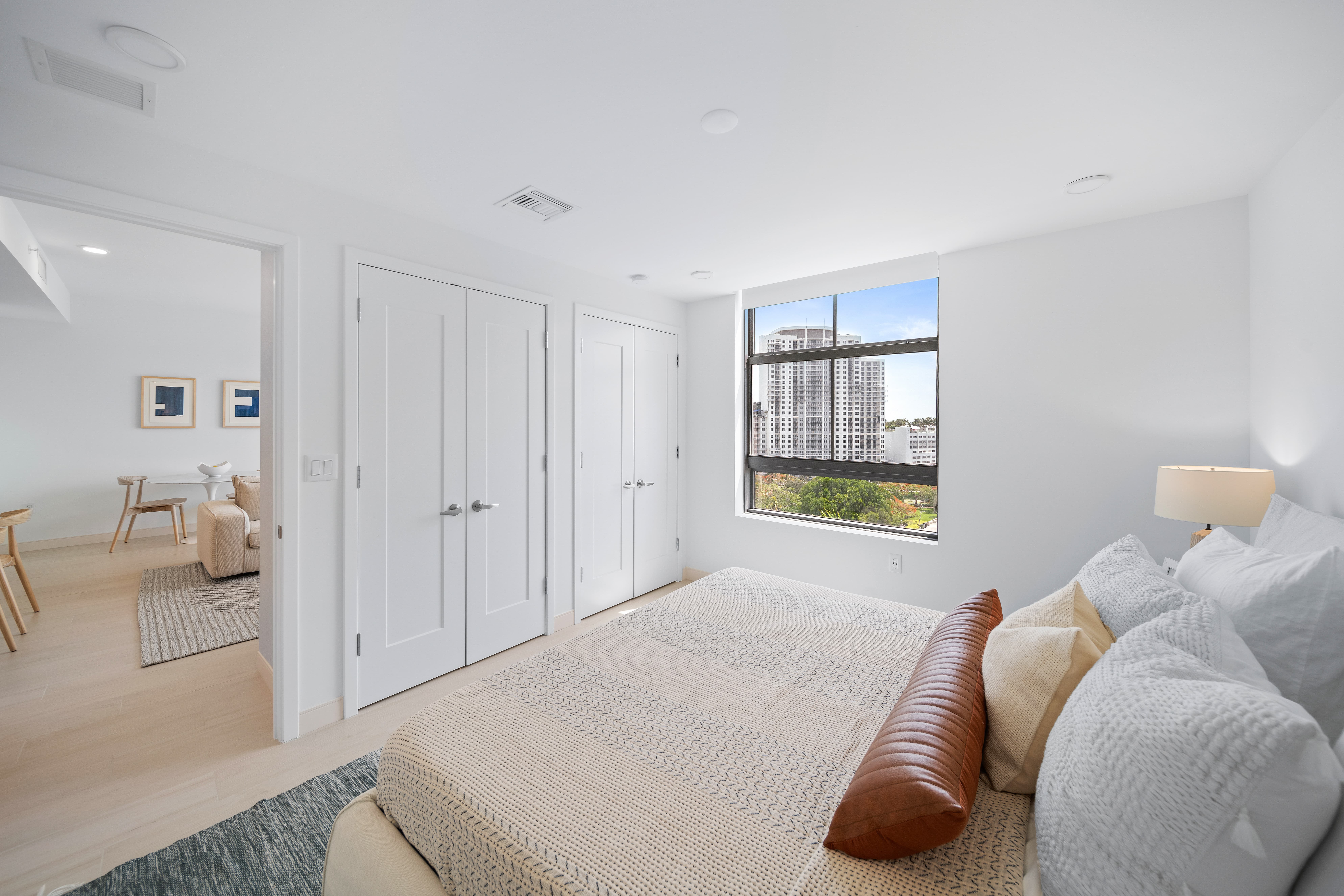 A modern bedroom featuring a neatly made bed with decorative pillows and a brown throw pillow. There are two white closets and a large window showing a city view. The room has light-colored walls and wooden floors.