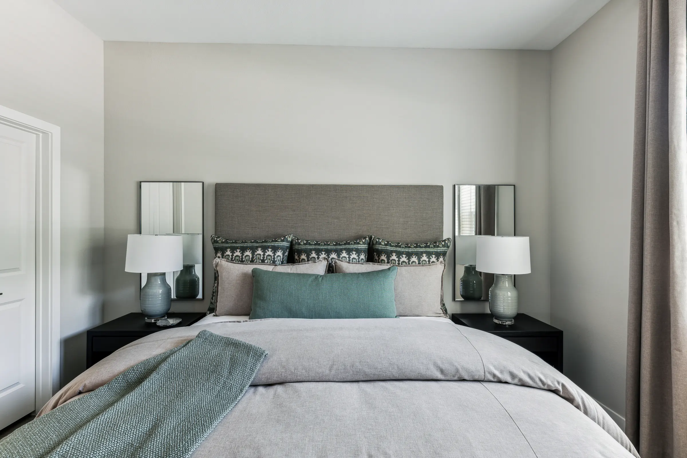 Elegant Modern Bedroom Design A modern bedroom featuring a neatly made bed with a grey comforter, decorative pillows, and a green throw blanket. There are two matching bedside tables with turquoise lamps and mirrors flanking the bed.