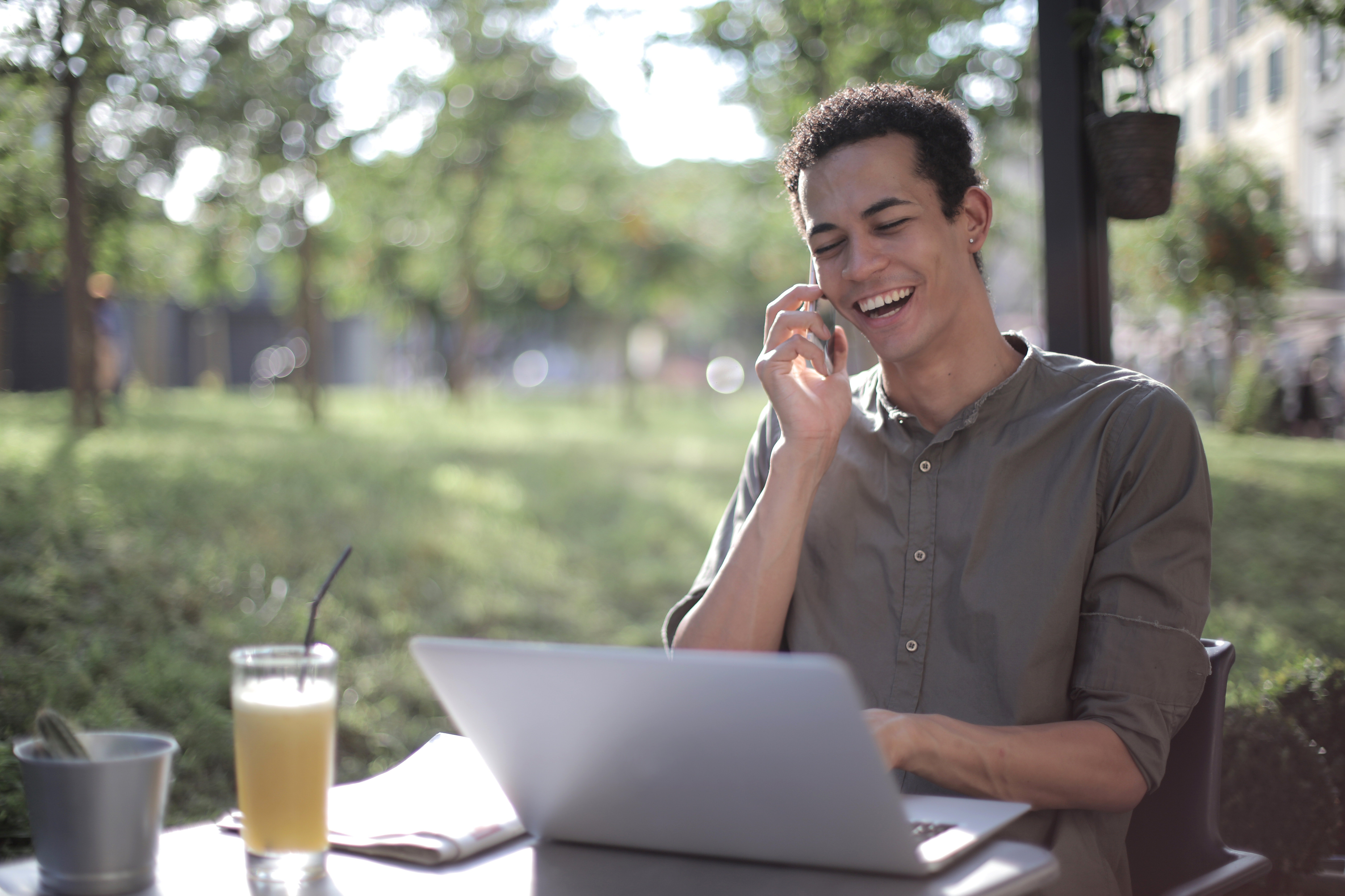 A young man sitting outside at a table with a laptop, smiling while talking on the phone, with a glass of juice on the table.