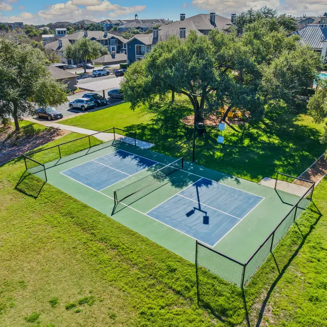 Aerial view of two tennis courts surrounded by grassy areas and trees, with a parking lot and residential buildings in the background.