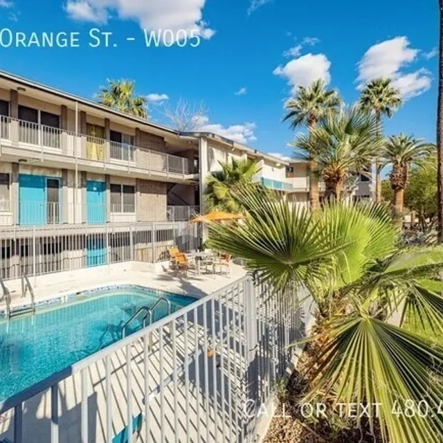 A view of an outdoor area featuring a swimming pool, palm trees, and a multi-story building with balconies. The pool area includes lounge chairs and is surrounded by a fence.