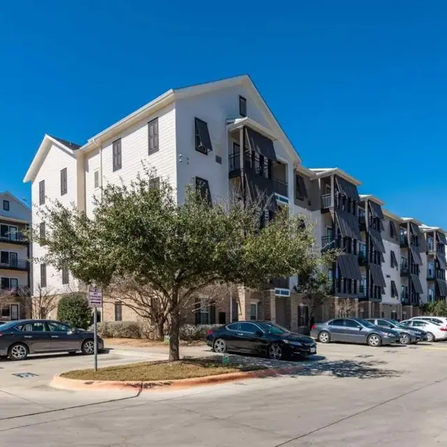 A modern apartment complex featuring multiple buildings with white exteriors and black shutters, set against a clear blue sky. Several parked cars can be seen in the foreground along a paved pathway lined with grass and small trees.