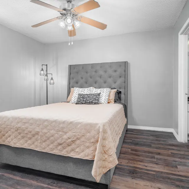 A modern bedroom featuring a gray upholstered bed with decorative pillows, a light brown quilt, and a ceiling fan. There are two lamps beside the bed and a doorway leading to another room in the background.