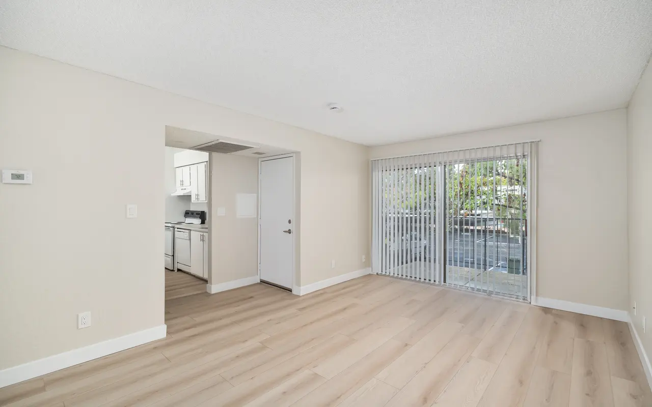 A bright and spacious living room with light-colored wooden floors, white walls, and a sliding glass door that leads to an outside area. Part of a kitchen is visible on the left side.