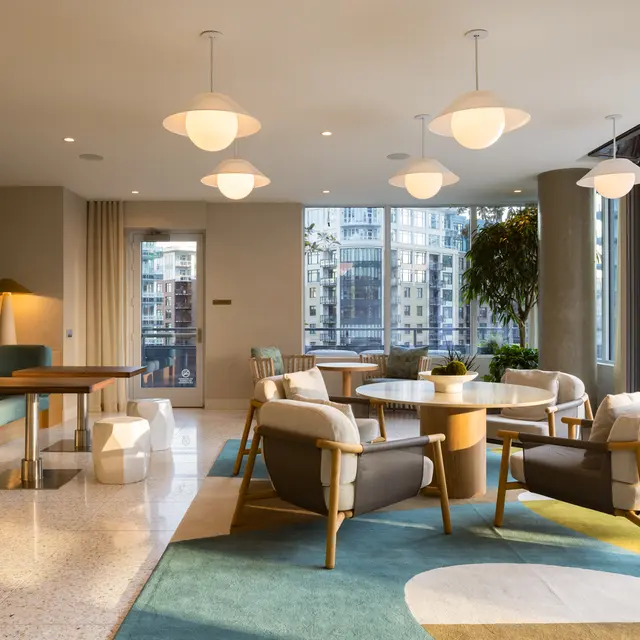A modern lounge area with elegant furniture and large windows, featuring a mix of comfortable seating, round tables, and potted plants, creating a bright and inviting atmosphere.