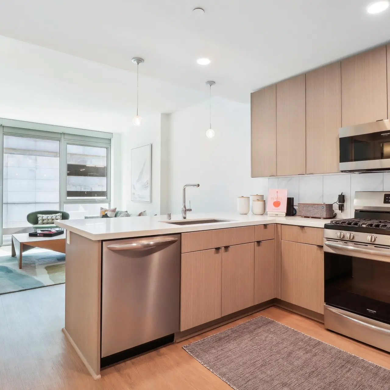 A modern kitchen with wooden cabinetry, stainless steel appliances, and a spacious layout. The kitchen includes a sink, stove, and dishwasher. There are pendant lights hanging above the kitchen island, and a large window allows natural light to flood the space, which is connected to a living area visible in the background.