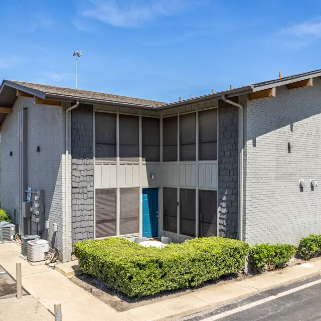 Exterior view of a two-story building with a grey facade and blue door, surrounded by greenery and a parking area.