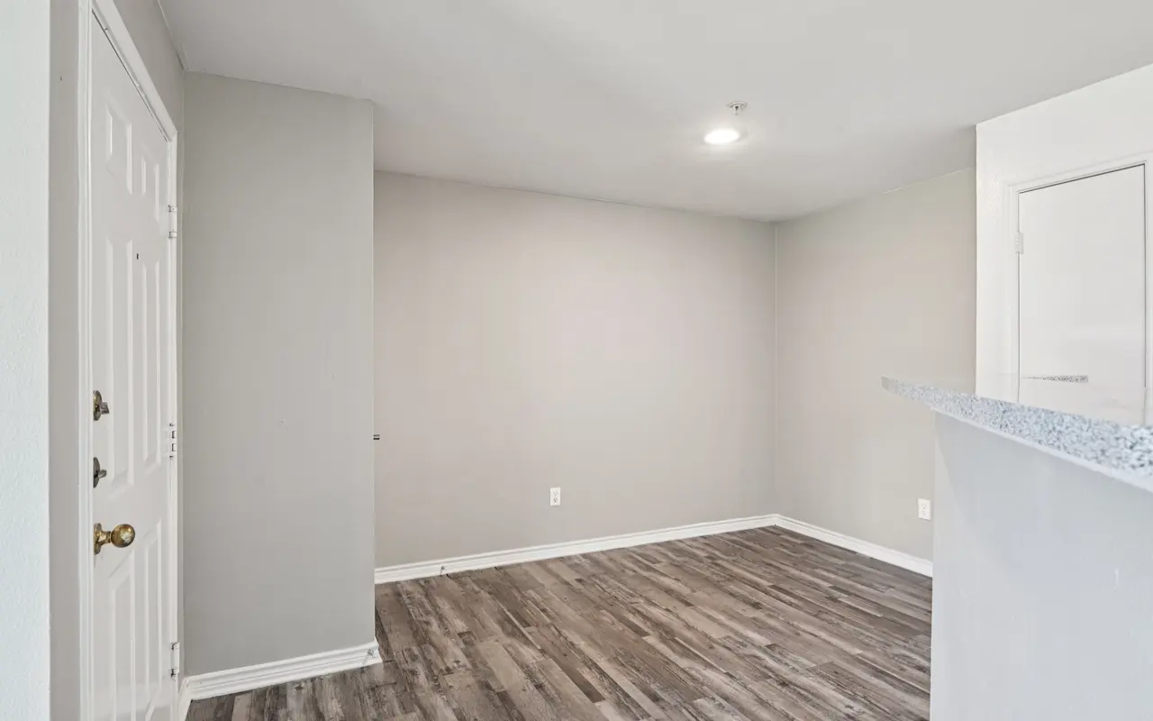 A spacious, empty room featuring light gray walls and wooden flooring. A doorway is visible on the left, and a small counter provides a division to another area.