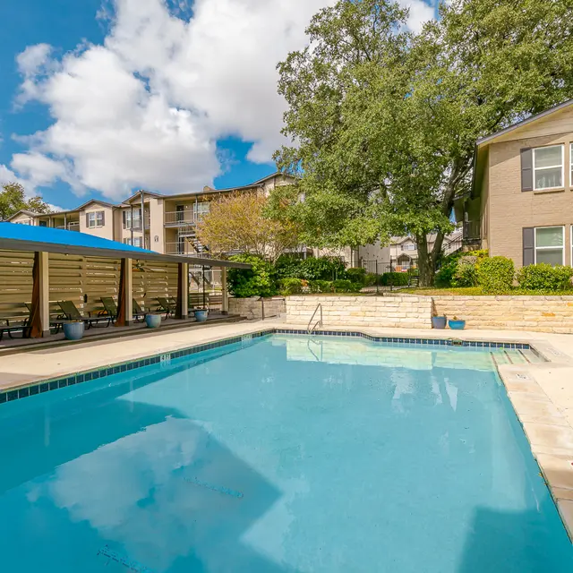 A clear swimming pool surrounded by lounge chairs and green landscaping, with apartment buildings in the background under a partly cloudy sky.