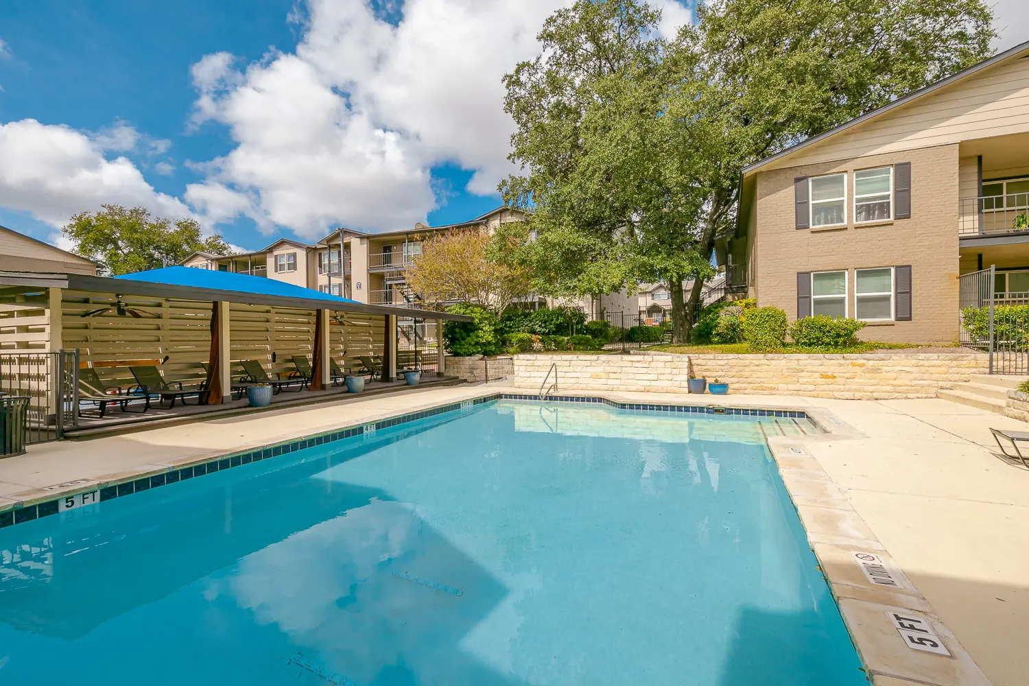 A clear swimming pool surrounded by lounge chairs and green landscaping, with apartment buildings in the background under a partly cloudy sky.
