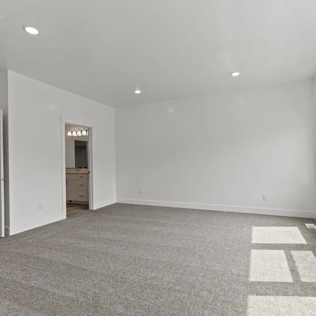 A spacious and bright living room with gray carpet, large windows, and white walls. There is an entrance leading to another room on the left side and natural light coming through the windows.