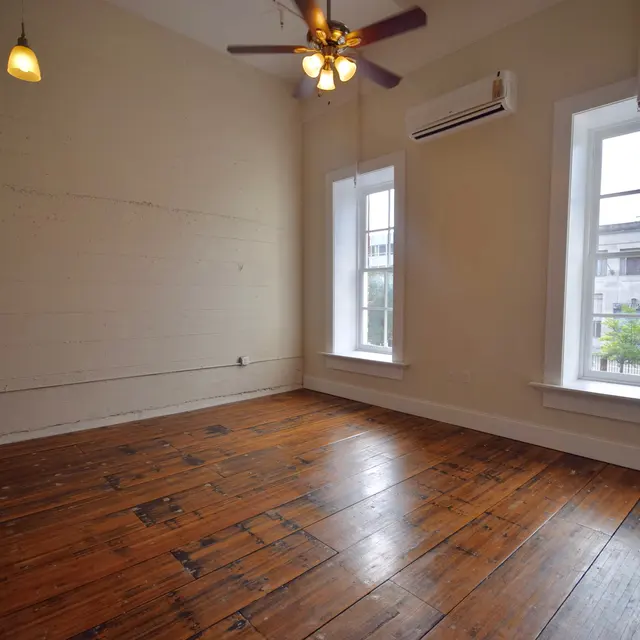 An empty room with wooden flooring, two windows on one wall, and a ceiling fan with three blades. There are two small light fixtures hanging from the ceiling.