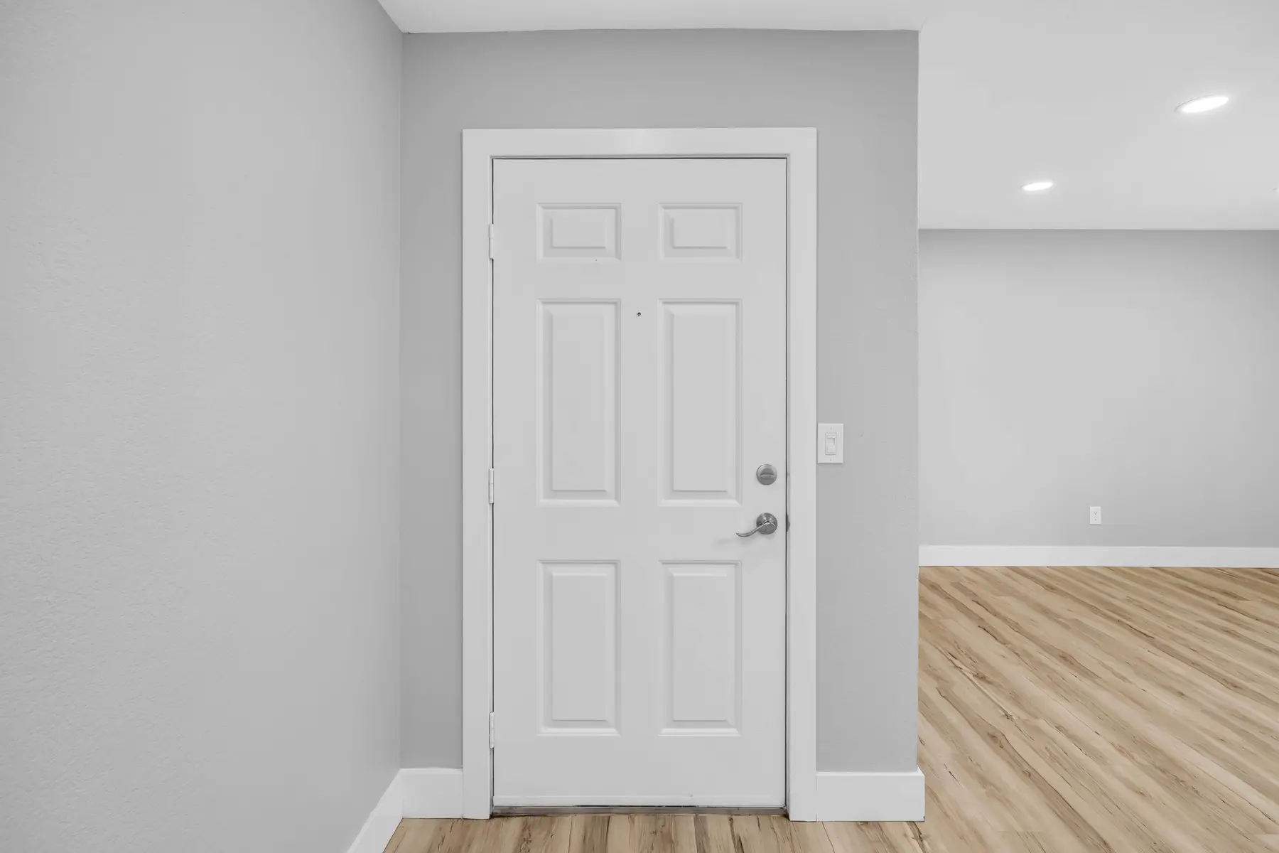 A modern entryway featuring a white front door next to a smooth gray wall. The floor is made of light-colored wood, providing a warm contrast to the walls. Bright overhead lighting adds clarity to the space.
