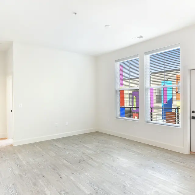 Spacious Living Room with Colorful Views An empty living room featuring white walls, large windows with blinds, and light-colored flooring. A door leads to an outdoor area, with colorful elements visible through the windows.