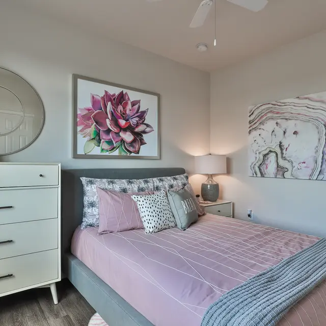 A cozy bedroom featuring a gray bed with pink bedding, decorative pillows, and a wall art piece. There is a white dresser beside the bed, a round mirror on the wall, and light-colored walls with three paintings.