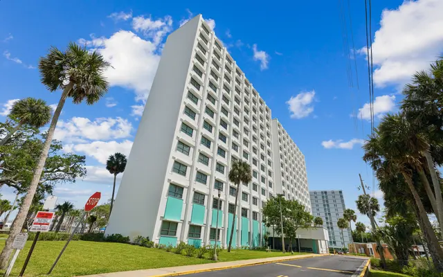 A modern hotel building with a white facade and teal accents on a sunny day, surrounded by palm trees and blue skies.