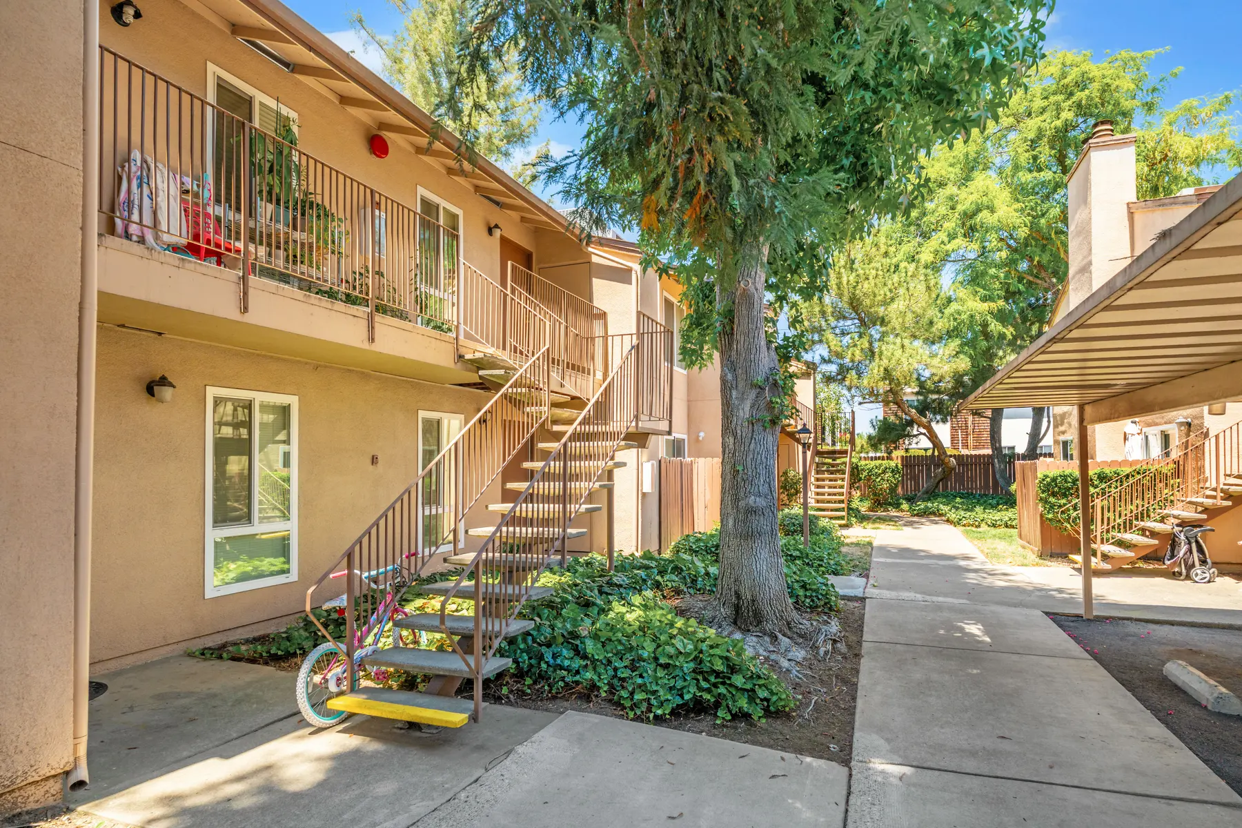 Exterior view of a two-story apartment complex with a staircase, surrounded by greenery and a pathway.