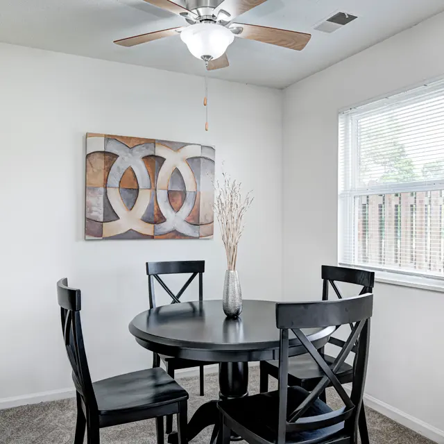 A small dining area featuring a round black table with four chairs, a decorative wall art piece, and a ceiling fan. There's a window letting in natural light.